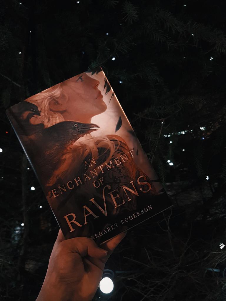 An Enchantment of Ravens by Margaret Rogerson held up in front of a dark background with little white lights