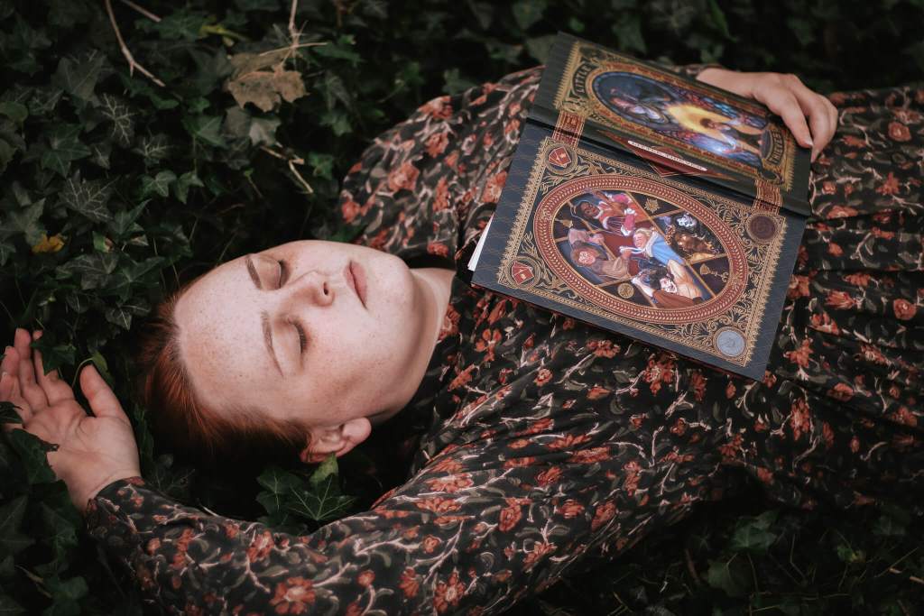 A girl with red hair and in a green dress with red flowers on it holds a detailed book on her chest called Little Thieves by Margaret Owen. The girl is lying in a copse of green leaves