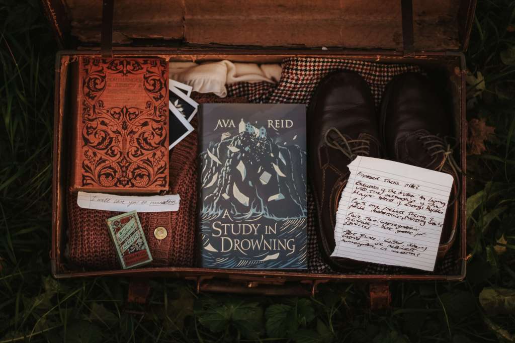 A Study in Drowning by Ava Reid sits in a suitcase that sits in a bed of grass. The suitcase holds a vintage books, polaroids, cigarettes, clothes, shoes.